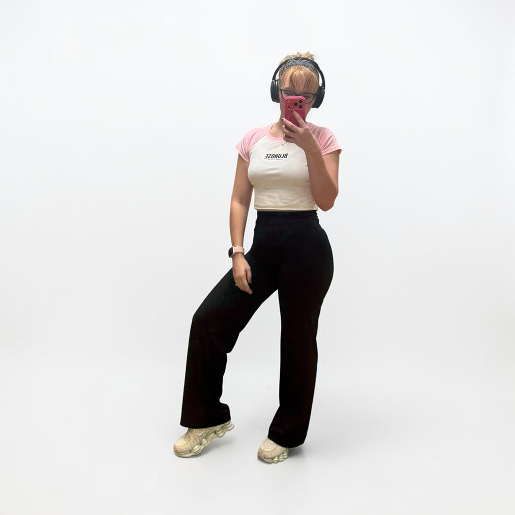 A woman posing in a studio setting wearing a short-sleeve crop top with a pink and cream color-blocked design, black wide-leg pants, and beige sneakers. She is holding a pink smartphone to take a mirror selfie while wearing black over-ear headphones.